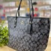 Cartera COACH tote en tono marrón oscuro con detalles negros y dorados.