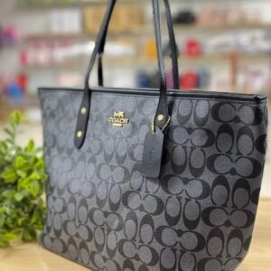 Cartera COACH tote en tono marrón oscuro con detalles negros y dorados.