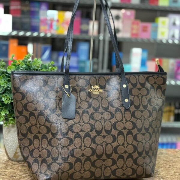 Cartera COACH tote en tono marrón oscuro con detalles negros y dorados.